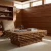 Wood-paneled living room with built-in shelves, a woven coffee table sculpture, and stacked books beside a cushioned chair bathed in natural light through high windows.