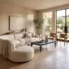 Soft beige living room with a large curved sofa, throw blanket, and modern abstract wall art near a glass door and windows opening to a garden.