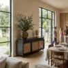 Interior dining area with a tall eucalyptus plant on a dark cabinet beside large floor-to-ceiling windows, table set for a meal nearby.