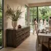 Tall potted olive tree on a carved wooden sideboard inside a bright dining area; large glass doors open to a green garden with guests outside.