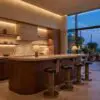 Warm wood kitchen with a long island and row of metallic bar stools, soft under-cabinet lighting, and a large window showing evening blue sky and string lights outside.