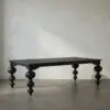 A large dark wooden table with thick, rounded legs standing in a bare room with a textured off-white wall behind it.