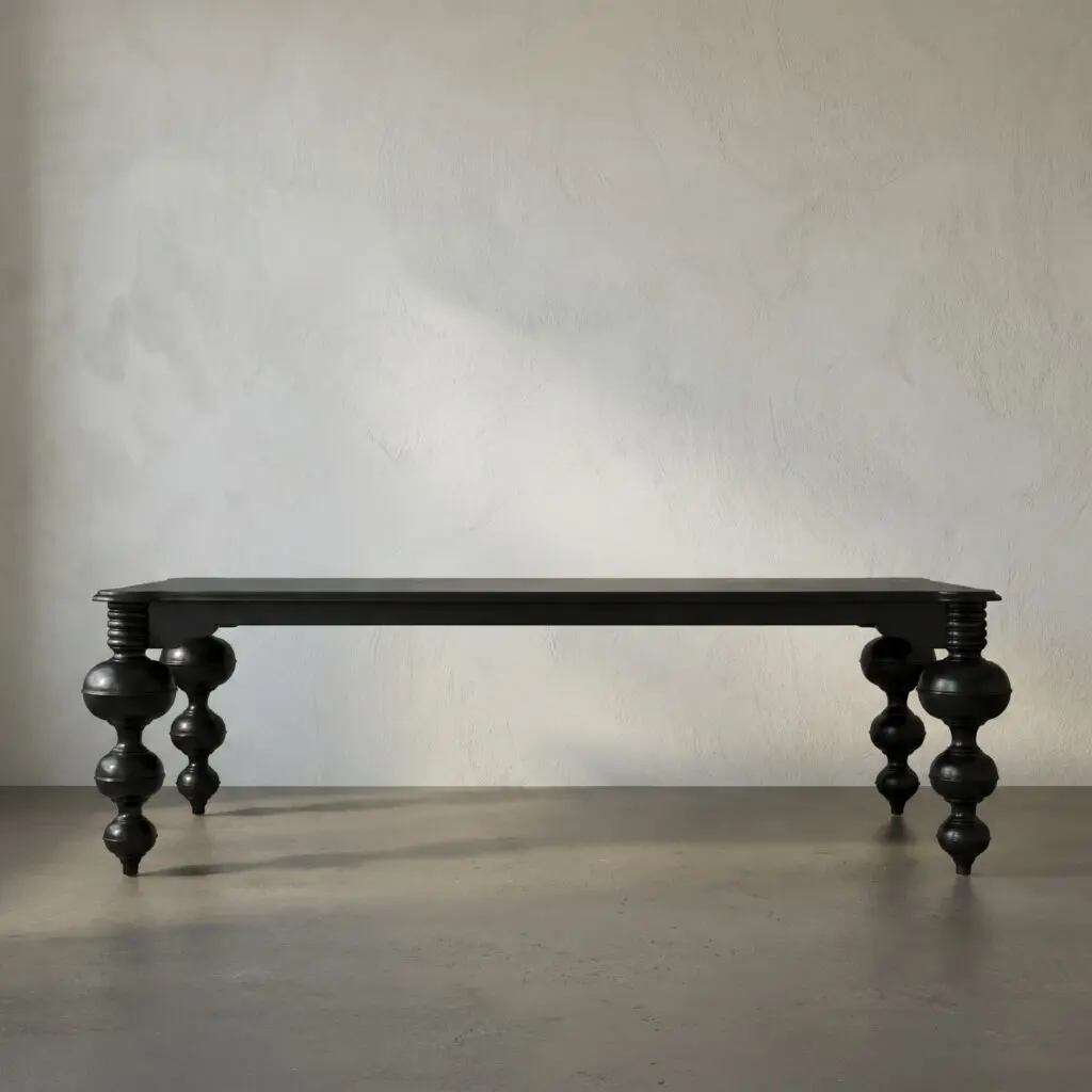 Long dark wooden table with thick, turned legs against a textured gray wall and concrete floor in a minimal room.