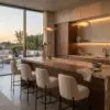 Modern kitchen with a long wooden island and cream boucle bar stools, warm pendant lights, and an outdoor seating area visible through large windows at sunset.