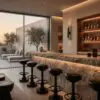 Modern bar area with a marble counter and black stools, glass wall showing trees outside, warm lighting in background.