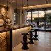Modern dining area with a long wooden bar, stone wall, and hanging pendant lights, large windows showing sunset outside.