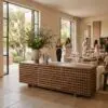 Open dining room with a sculptural wooden cabinet as a centerpiece, a tall leafy arrangement, and large windows letting in daylight with guests in the background.