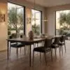 Sunlit dining area with a long wooden table, black leather chairs, and warm pendant lights; large windows view trees outside.
