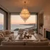 Living room at sunset with a beige sofa, coffee table, and a tiered crystal chandelier; ocean view through glass doors outside.