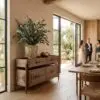 Wooden sideboard with a geometric pattern, a large potted eucalyptus plant, and glass doors letting in natural light; blurred guests in background.