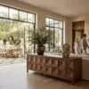 Large wooden cabinet with a diamond-patterned front in a bright living space, next to expansive glass doors and a tall leafy plant on top or nearby.