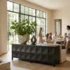 Tall indoor plant in a textured stone planter sits on a dark geometric cabinet by large glass windows, with people dining in the background.