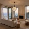 Modern living room with beige sofa, glass doors, and a crystal chandelier overlooking the ocean at sunset.