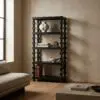 Tall black decorative bookshelf in a corner holding a white ceramic vase, small house figurines, and a stack of books against a beige textured wall near a window.