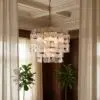 Elegant chandelier with frosted glass panels hanging from a coffered ceiling, framed by beige curtains and leafy plants nearby.
