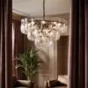 Elegant glass chandelier with textured, circular panels hanging from a chain, glowing softly in a living room framed by burgundy curtains and plants.
