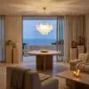 Living room with beige seating, round wooden table, candle centerpiece, and ocean view through sliding glass doors.