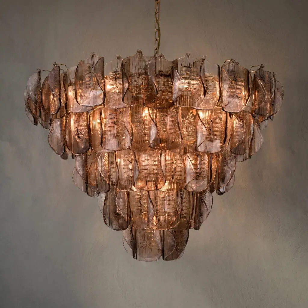 Copper-toned glass chandelier with layered petal-like prisms hanging from a chain, lit warmly against a neutral wall.