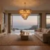 Modern living room with a sculptural chandelier and an ocean view through open glass doors; warm lighting, neutral furniture, and stone wall on left.