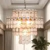 Elegant gold chandelier made of overlapping circular pink glass discs hanging from a ceiling in a bright room with draped curtains and plants nearby.