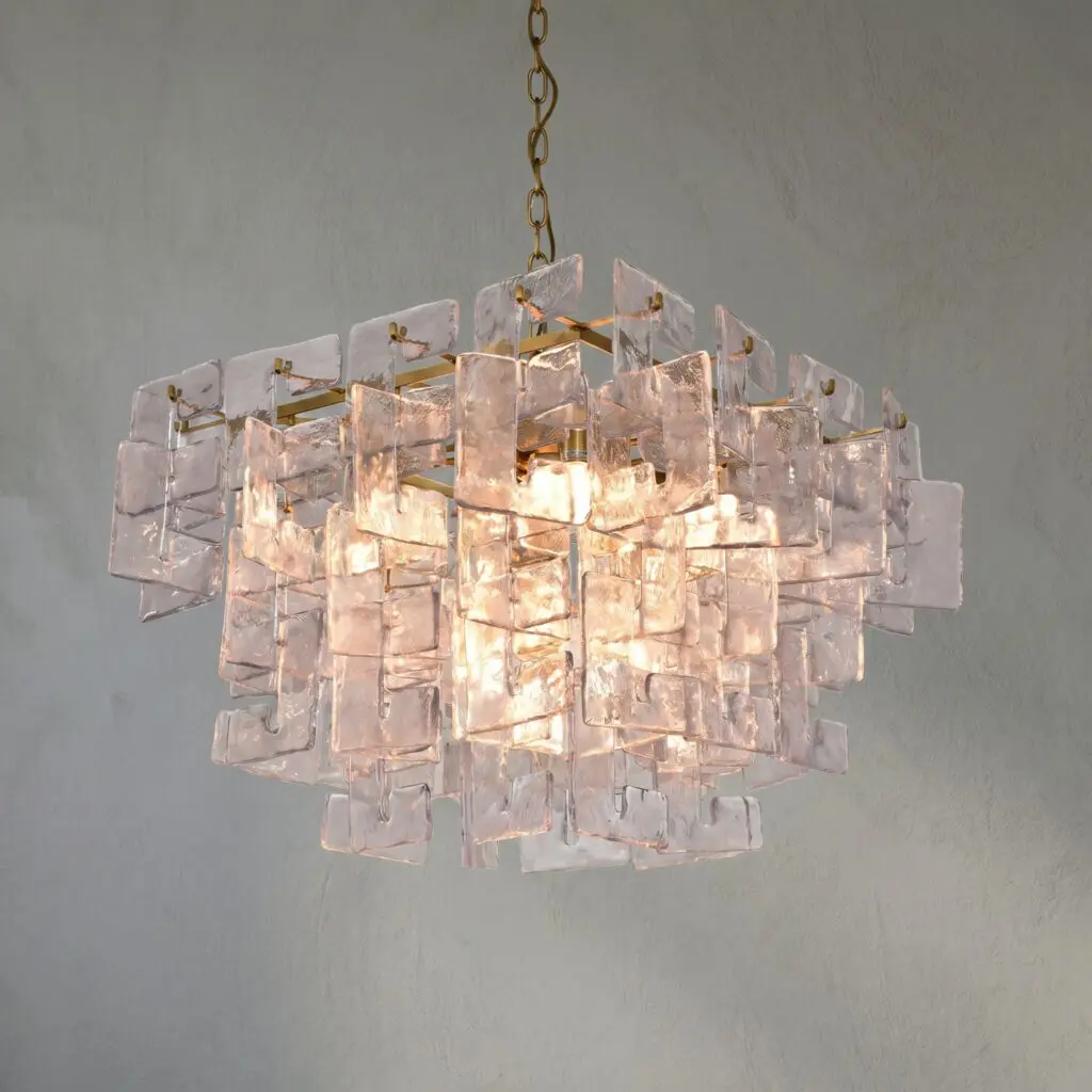 Chandelier made of glass blocks hanging from a brass chain against a gray wall, softly lit from within.