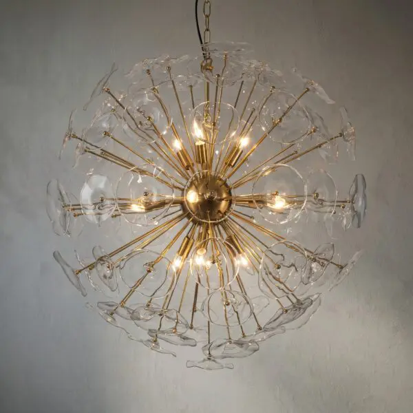 Crystal glass and gold chandelier with numerous glass petals radiating outward, lit against a gray wall.