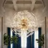 Ornate glass and gold chandelier hanging from a decorative coffered ceiling, with blue velvet curtains and green plants nearby.