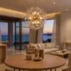 Circular wooden dining table with ceramic bowls and a globe-like glass chandelier, overlooking the sea at sunset in a modern living room.