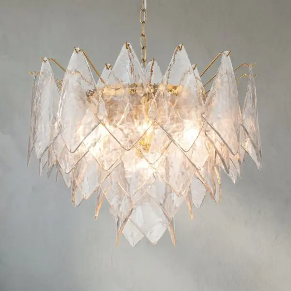 Crystal chandelier with clear glass prisms and gold metal accents hanging from a chain against a gray wall.