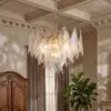 Crystal chandelier with faceted glass prisms hangs from a chain in a room with decorative crown molding and beige curtains, plus a potted plant nearby.