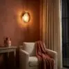 Orange-walled seating area with a glowing wall lamp beside a beige armchair and rust-colored throw, and a ceramic pitcher on a wooden table by the window.