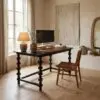 Desk workspace by a sunlit window with a black monitor, ceramic vases, and a large beige abstract textile hanging on the wall behind it.