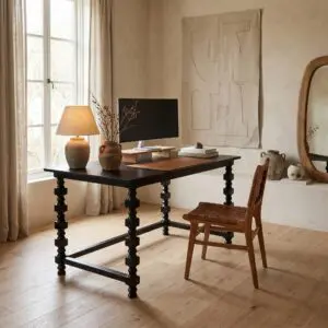 Desk workspace by a sunlit window with a black monitor, ceramic vases, and a large beige abstract textile hanging on the wall behind it.