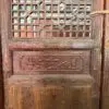 Old wooden door with a geometric lattice window and a carved relief panel below, showing an aged, reddish-brown finish.