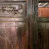 Close-up of an antique carved wooden door with a floral relief panel on the left, dark weathered wood, and a glimpse of brick wall through the right doorway.
