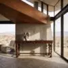 Interior scene with a diagonal wooden beam, concrete column, and floor-to-ceiling glass walls overlooking a desert landscape; a sculpture bust sits on a wooden console with books nearby.