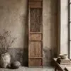 Tall carved wooden cabinet against a weathered plaster wall, with a ceramic vase of bare branches to the left and bowls on a wooden table to the right.