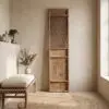 Tall wooden cabinet with a lattice panel stands against a beige wall in a minimalist room, with a round woven rug nearby and a plant on the left shelf.