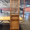 Tall antique wooden cabinet with a woven lattice door panel, standing on a wooden table in an empty industrial warehouse.