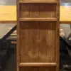 Tall, weathered wooden cabinet with a decorative lattice panel on top, placed on a wooden platform in a warehouse or workshop setting with tables in the background