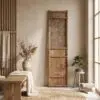Wooden armoire with geometric lattice panel in a neutral, minimalist room; dried grasses in a vase on a bench nearby.
