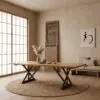 Wooden table with black X-shaped legs on a round woven rug in a minimalist beige room with soft light from large windows.