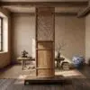 Tall wooden screen with a woven lattice panel stands centrally in a sunlit, rustic room; a table with a small sculpture and books sits to the left, while a large blue-and-white ceramic vase and a bare-branch arrangement sit to the right.