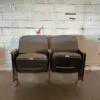 Two old, black theater seats with wooden armrests sit side by side in front of a rough, white-painted brick wall.