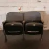 Two adjacent, upholstered theater seats with worn Armrests, set against a rough white brick wall.