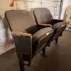 Two dusty theater chairs with wooden armrests sit side by side against a white, worn brick wall in an industrial space.