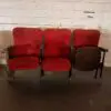 Row of three red auditorium chairs with metal frames against a white brick wall; the rightmost chair is missing its back panel.