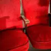 Pair of red upholstered theater seats with a wooden armrest between them, worn fabric against a white wall.
