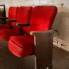 Row of red upholstered theater seats with wooden armrests against a white brick wall.