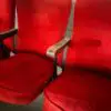 Red velvet theater seats with metal armrests, arranged in a row against a light brick wall.